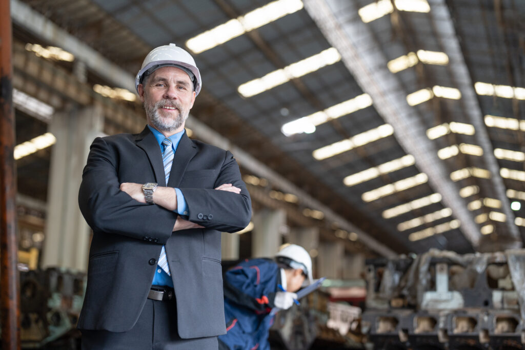 Senior businessman wearing helmet safety standing arms crossed in heavy metal factory industrial. In behind Asian engineer woman is checking machine equipment parts. Work success concept.